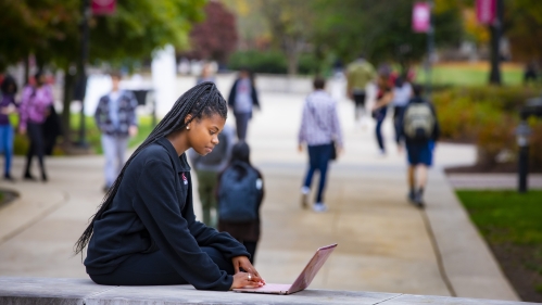 Student working on laptop outside while others walk by in background.