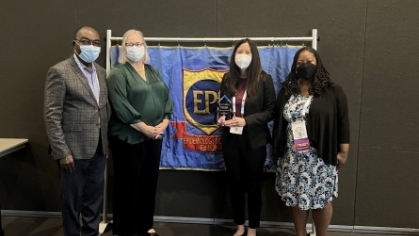 Photo of four people in masks in front of sign holding an award.