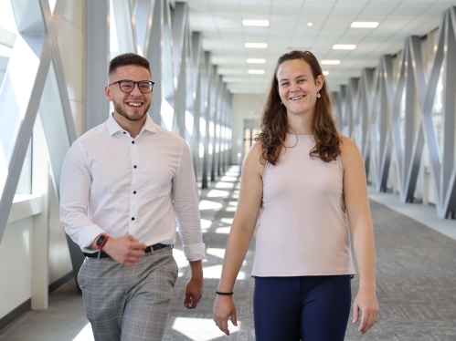 Two people walking in hallway and smiling at camera. 