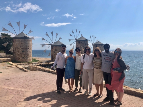 Six people standing outside in front of windmills and ocean.